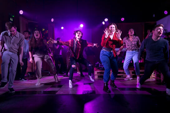 Production image : A large group of young people, facing the audience, mid dance or shout or fight. They are full of energy and passion, taking up space.