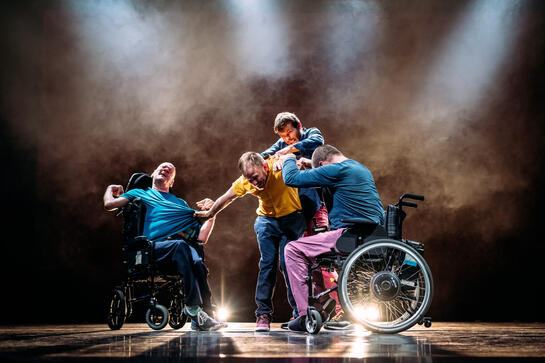 Production image : A struggle between four men, two standing, two using wheelchairs. T-shirts are pulled, fists and arms are locked. They are back-lit, highlighting the angles their bodies are making.