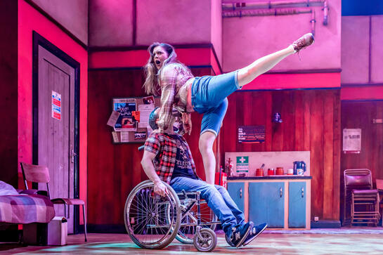 Production image : A woman stands in arabesque on the lap of a man who uses a wheelchair. Everyone is bathed in pink with a fantastical quality.