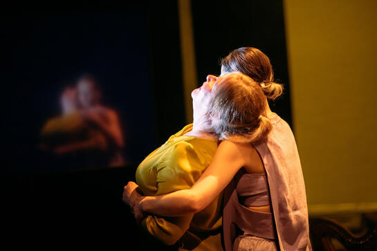 Production image : Two people in an embrace. A woman leans her head back on to the other's shoulder. A vulnerability. Soft fabrics and lighting.