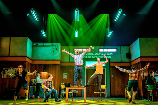 Production image : An ensemble cast of 7 people mid song. Some stand on chairs and tables and there's a guy at a piano. Arms are out stretched, big movements under strong green spotlights. The set suggests a village hall and we can see projected captions w