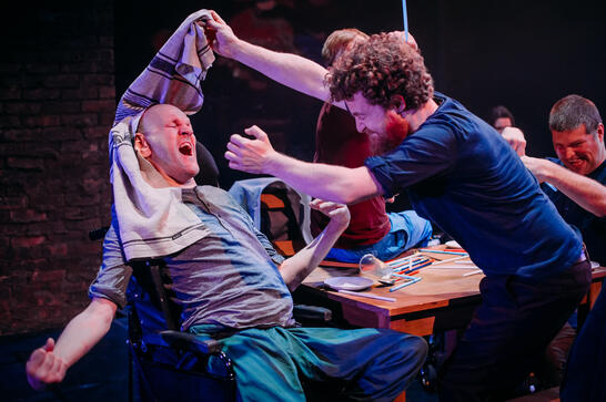 Production image : Two men are mucking around with a teatowel - one, a wheelchair user, is trying to escape his head being covered.