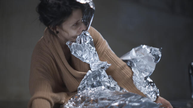 Film image : A woman is half covered in tinfoil. In her mouth, under her arm. Its shiny texture contrasts her muted sand coloured jumper.