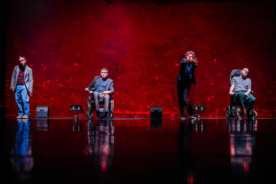 Production image : A deep red textured back wall. Four men waiting at the back of the stage, all facing us, the audience. Two wheelchair uses and two standing.