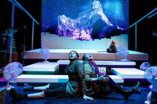 Production image : A man and a woman sit back-to-back in the centre of the image, both looking upwards with mouths open. A winter mountain backdrop behind them and various white platforms making steps. They are wearing an unusual collections of tweed short