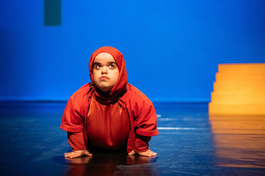 A woman lies on the ground, hand in front and pushes her head and shoulders up. Her eyes search upwards. She is wearing all amber coloured clothes and headscarf.