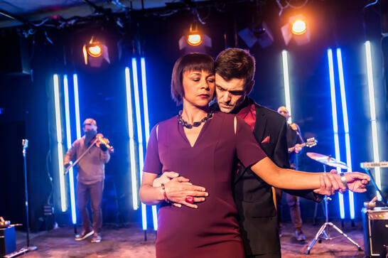 A man and a woman in a slow dance embrace. Musicians in the background.