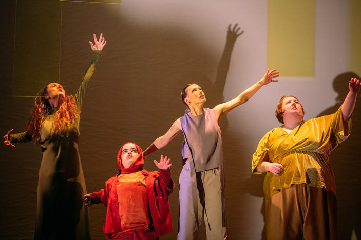 Production image : four women stand in a line across the stage. They each wear one colour; sage, amber, lilac, gold. Their left hands reach upwards and the heads and eyes follow. The woman dressed in amber wears a macthing headscarf.