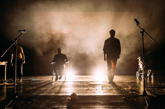 Production image : back-lit dominating bodies moving towards the audience. Some walking, some using wheelchairs. Smoke fills the space with microphones on stands close to us.