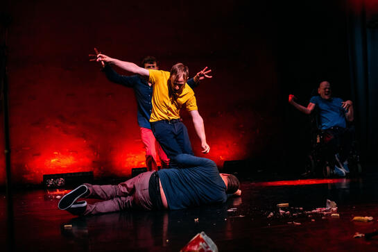 Production image : a man lies on the floor, on his side, head tucked in. Another stands above him, arms in the arms and weight into his feet; is he kicking or dancing? Two other men in the background, one a wheelchair user, also have their bodies and arms