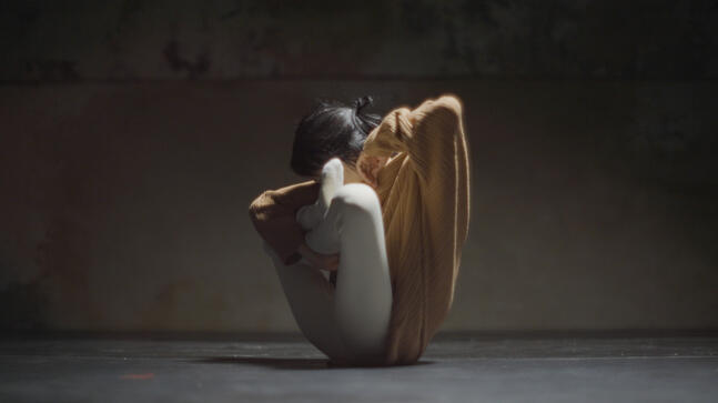 A film image : A female body on the floor, knees folded up towards head, elbows out. Stone and sand coloured clothes.
