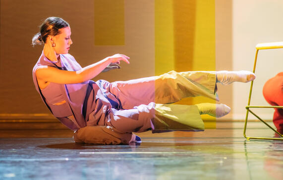 Production image : a woman balances on her pelvis on the floor, legs outstretched, bathed in yellow light.